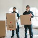 Two packers and movers carrying labelled cardboard boxes into a newly built house – shifting checklist by GharVistar